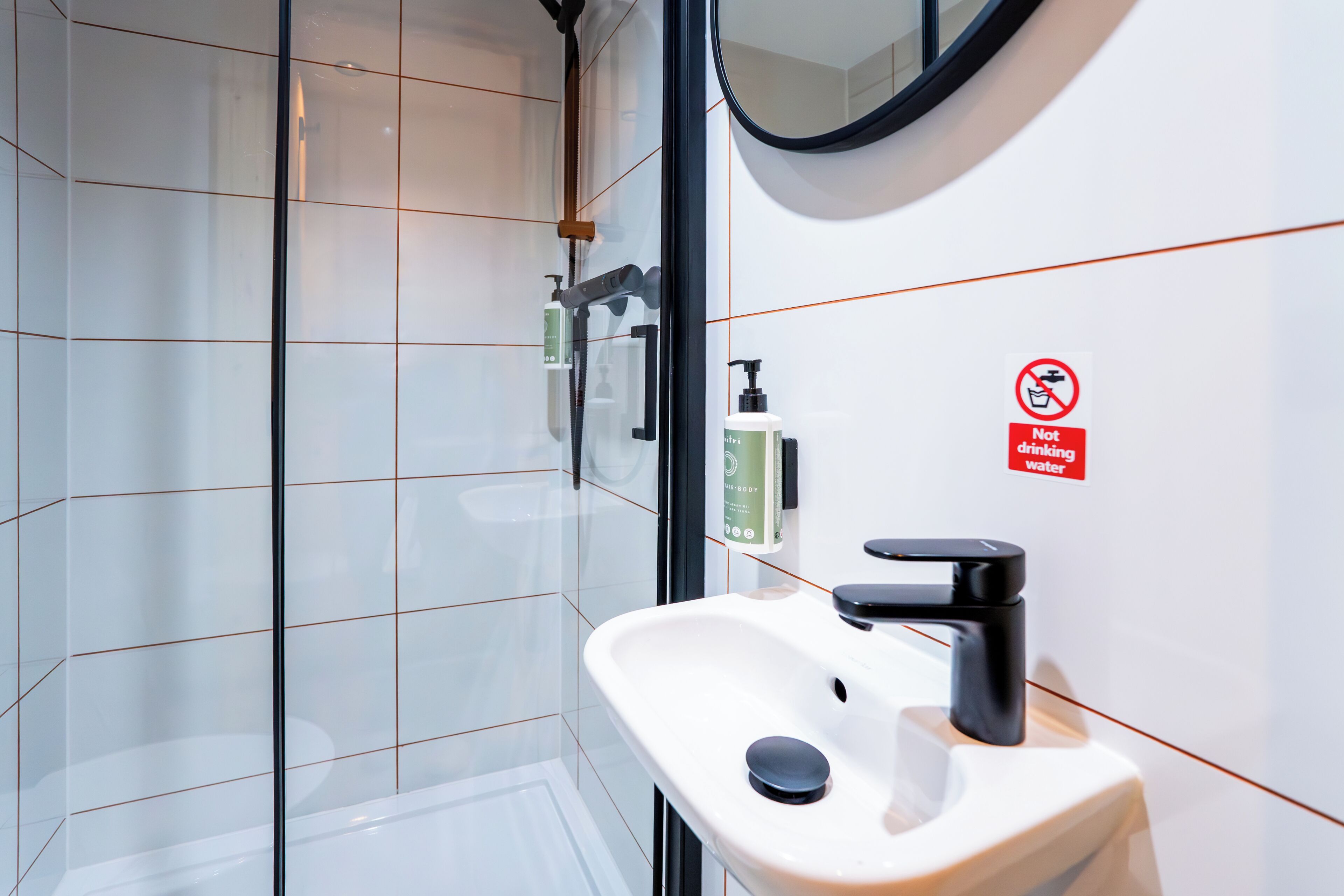 A modern bathroom with a glass shower enclosure, a white sink with a black faucet, and a 'Not drinking water' sign.