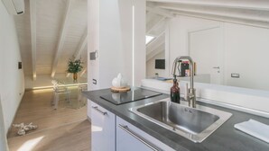 A modern kitchen with a large stainless steel sink, a glass dining table with chairs, and a ceiling with exposed beams. - Como Sky Suite by Rent All Como (Como)
