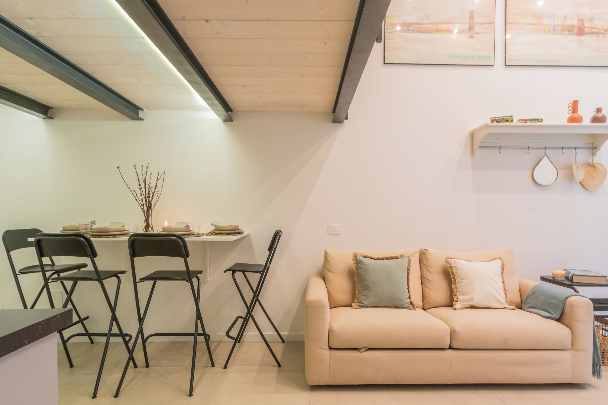 A modern living room with a beige sofa, black chairs, a dining table, and a kitchen area with a shelf.