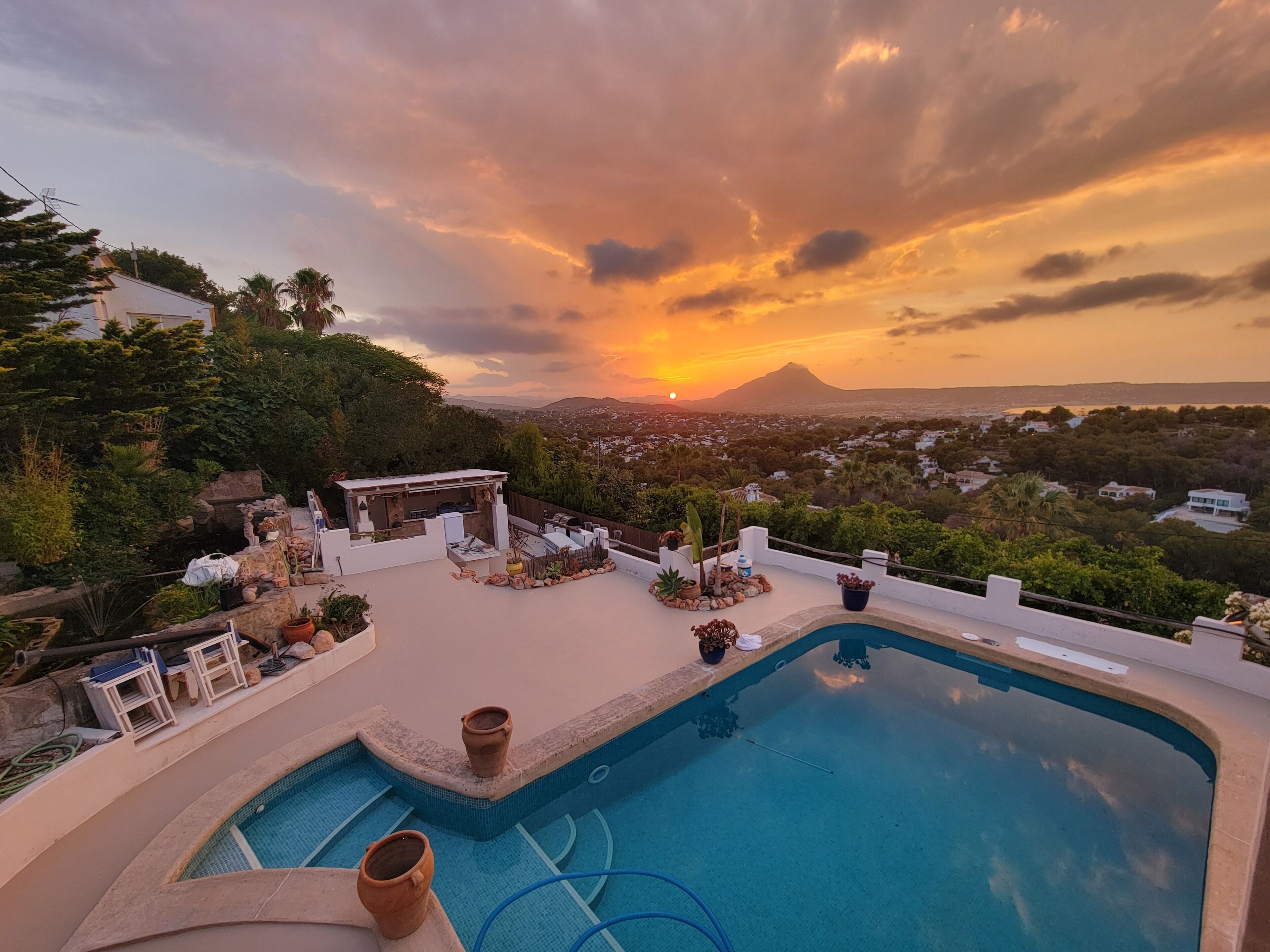 Ein Pool mit Pergola und Sitzbereich im Freien, umgeben von üppiger Vegetation und Blick auf eine Stadtlandschaft und einen Berg bei Sonnenuntergang.