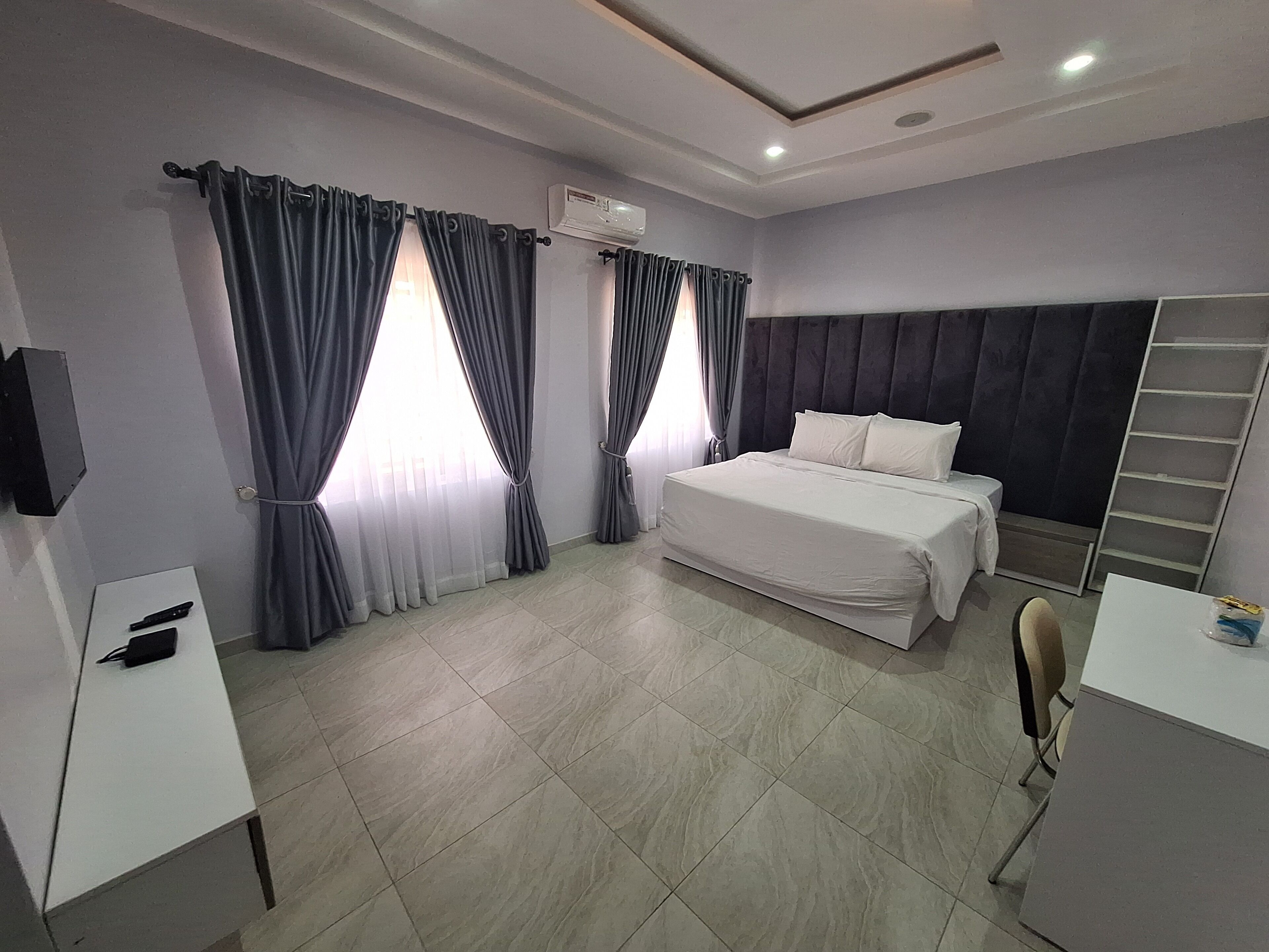 Une chambre d’hôtel moderne avec un grand lit, un bureau et une chaise. On y trouve une fenêtre avec des rideaux, un luminaire au plafond et un appareil de climatisation.