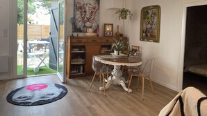 Un coin repas avec une table ronde, des chaises et un tapis décoratif. On y trouve un buffet en bois contenant de la vaisselle et une plante. Un tableau est accroché au mur, et un lustre est suspendu au plafond.