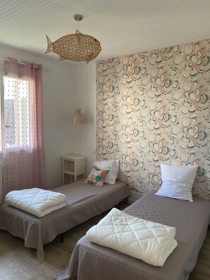 Habitació amb dos llits individuals, paper pintat amb un dibuix, llum penjant i una finestra amb cortines.