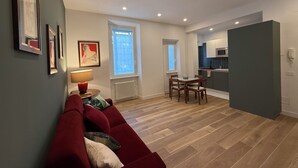Een woonkamer met een rode bank, een eettafel met stoelen, een keukenhoek en een raam met zonwering.