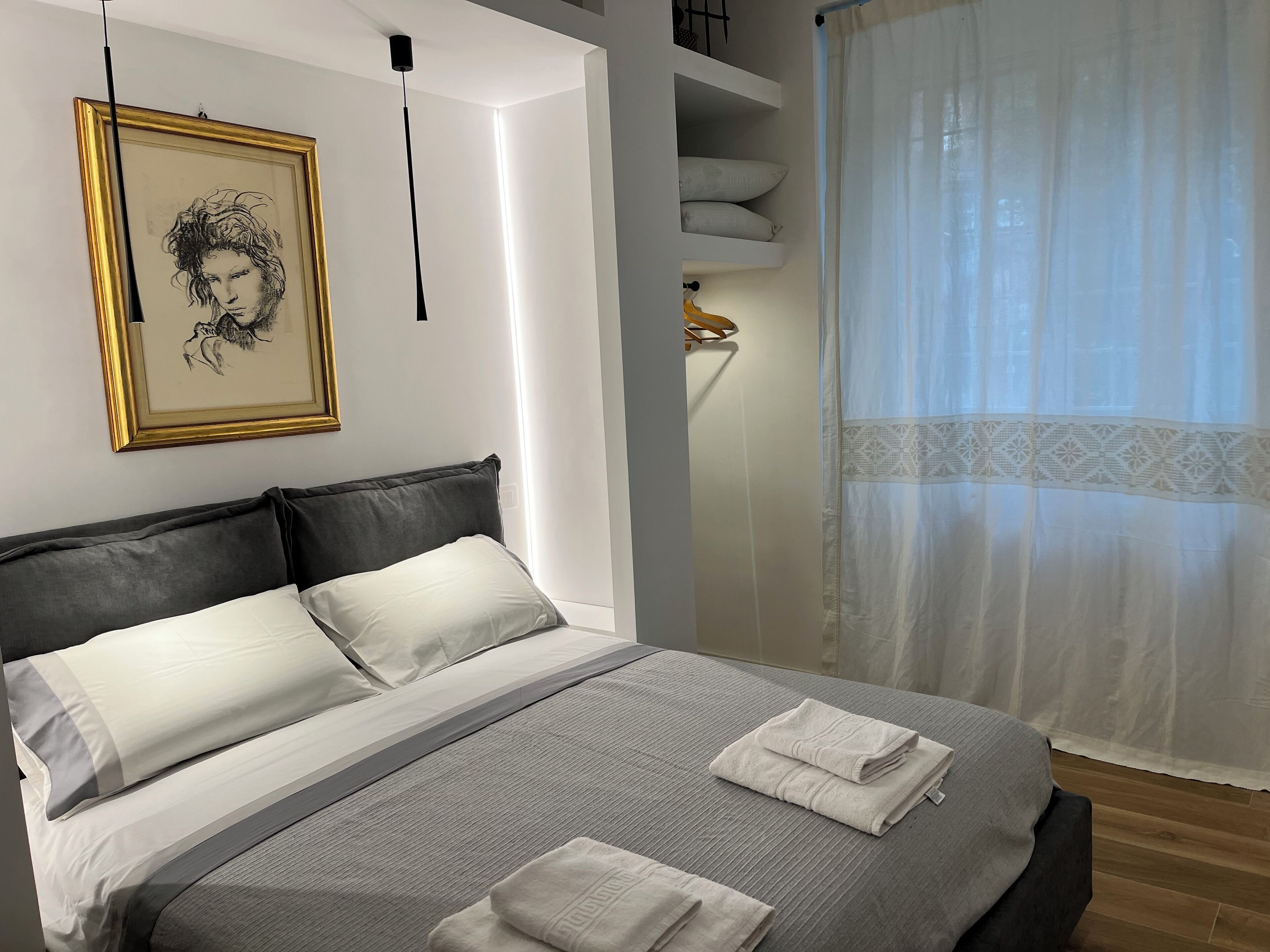 Un lit bien fait, agrémenté d’oreillers, une table de chevet avec une lampe, une œuvre d’art encadrée au mur et une fenêtre avec des rideaux légers.