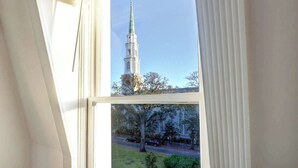 A window view of a tall white church tower with a cross on top, surrounded by trees and a road with cars. - 1872 Historic King Suite | Live Oak Views! (Savannah)