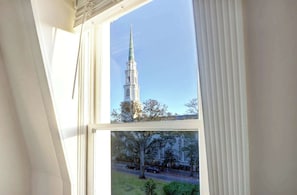 Blick aus dem Fenster auf einen hohen, weißen Kirchturm mit Kreuz, umgeben von Bäumen und einer Straße mit Autos.