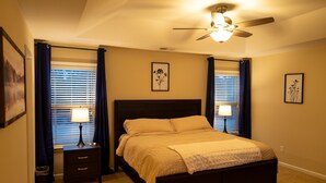 Une chambre à coucher comprenant un lit, deux tables de chevet avec des lampes, un ventilateur de plafond et deux fenêtres avec des rideaux.
