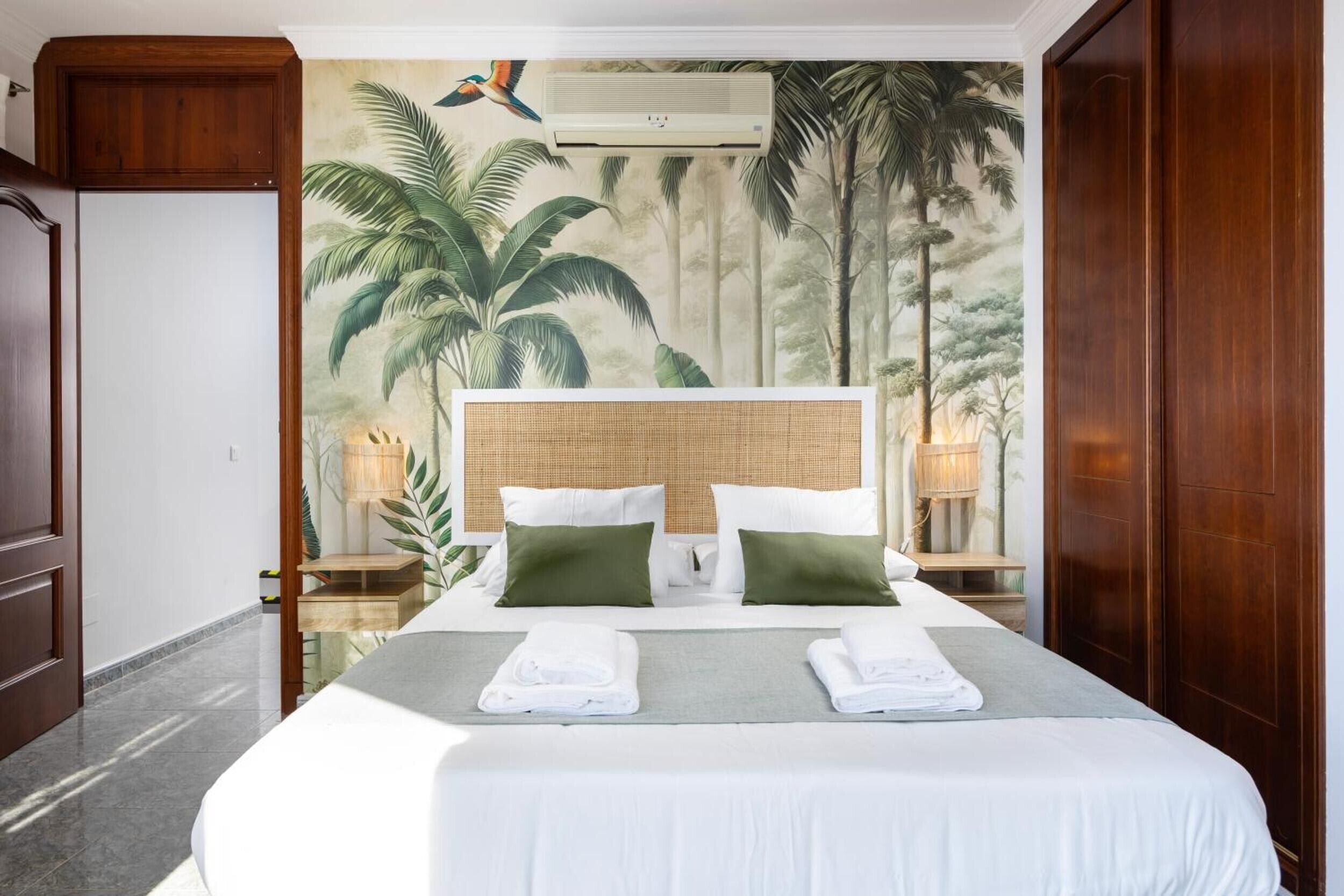 A hotel room with a large bed, a tropical mural, a wooden headboard, and a nightstand with a lamp.