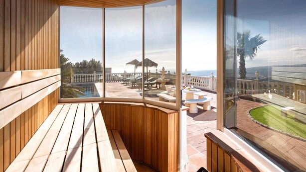 A wooden deck with a curved glass wall offering a view of an outdoor patio with tables and umbrellas. - Villa Selvatic with Sea Views, Pool, Sauna, and Cinema (Arenas)