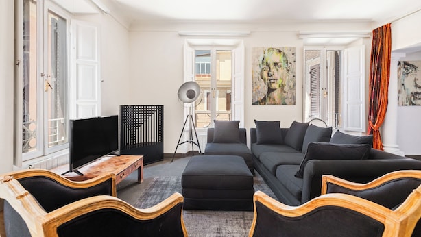 Un salon moderne avec un canapé, un pouf et des chaises en bois, un téléviseur et des œuvres d’art accrochées aux murs.