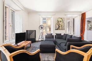 Ein modernes Wohnzimmer mit einer Couch, einem Hocker, Holzsesseln, einem Fernseher und Kunstwerken an den Wänden.