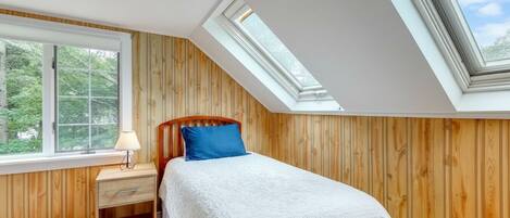 A bedroom with a sloped ceiling, wooden panel walls, a bed with white bedding and a blue pillow, a bedside table with a lamp, and a window with a view of trees.