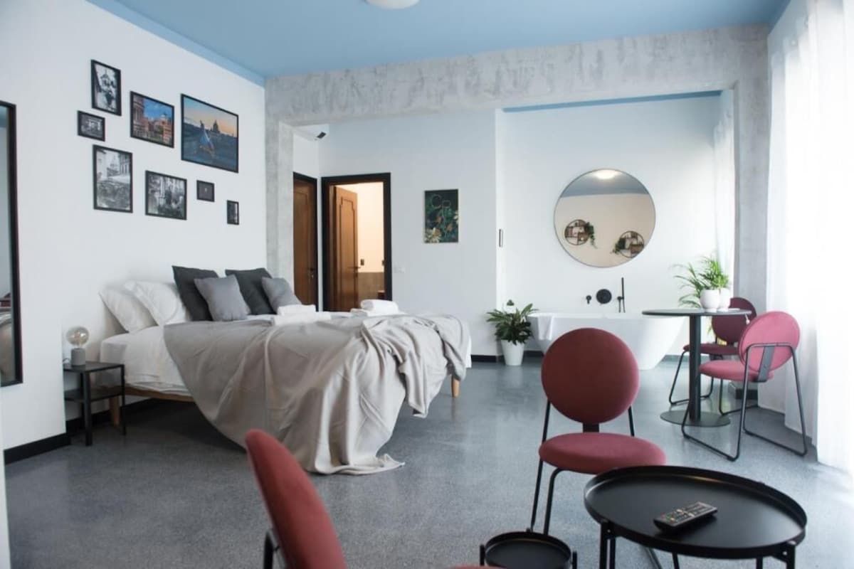 Moderni hotellihuone, jossa on sänky, työpöytä, tuoli, pyöreä peili ja pieni pöytä.