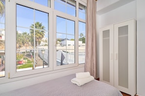 Une chambre avec une grande fenêtre, un lit recouvert d’une couverture blanche et une armoire.