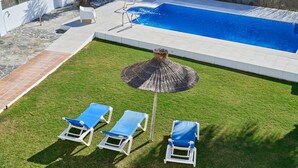Ein Schwimmbecken mit gefliestem Deck, einer Rasenfläche mit Liegestühlen und einem dekorativen Sonnenschirm.