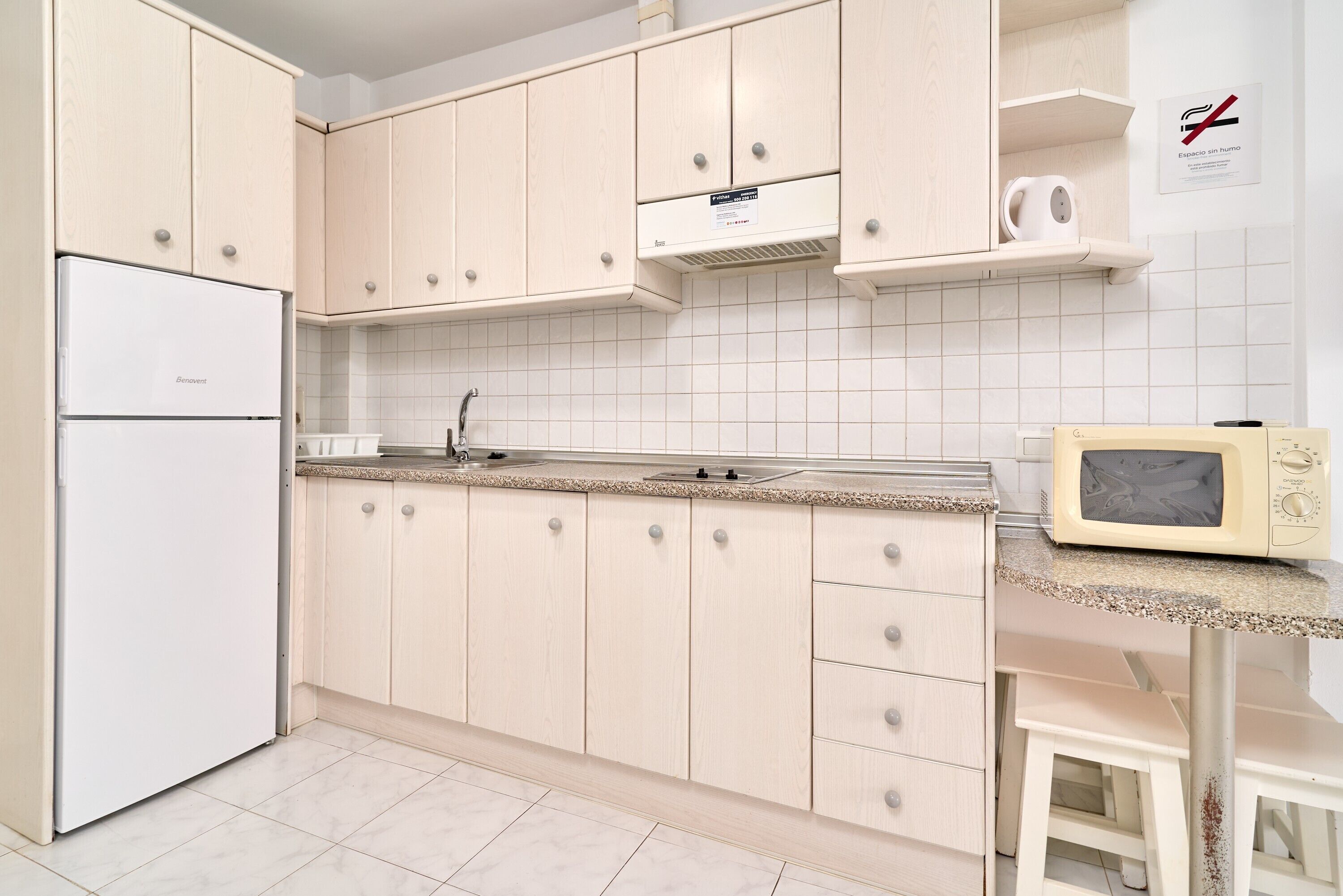 Una cocina con armarios de madera clara, un refrigerador blanco, un microondas y una salpicadura trasera de azulejos.