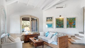 Un salon spacieux avec un canapé blanc, une table basse en bois et un fauteuil en osier. Trois œuvres d’art encadrées ornent le mur, et une fenêtre est dotée de volets.