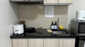 Eine kleine Küche mit hellen Holzschränken, einer schwarzen Arbeitsplatte, einer Edelstahl-Dunstabzugshaube, einer Kaffeemaschine, einem Wasserkocher, einem Spülbecken mit gelbem Seifenspender und einem kompakten Kühlschrank.