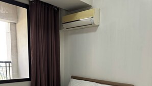 Ein ordentlich bezogenes Bett mit weißer Bettwäsche, einem braunen Kopfkissen und einem gefalteten braunen Handtuch. Eine Klimaanlage ist an der Wand über dem Bett angebracht.
