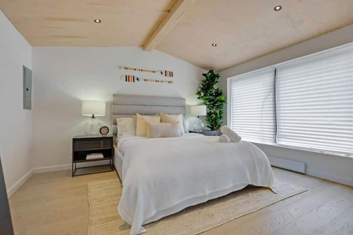 Un dormitorio con cama, mesitas de noche, lámpara, planta en maceta y ventana con persianas.