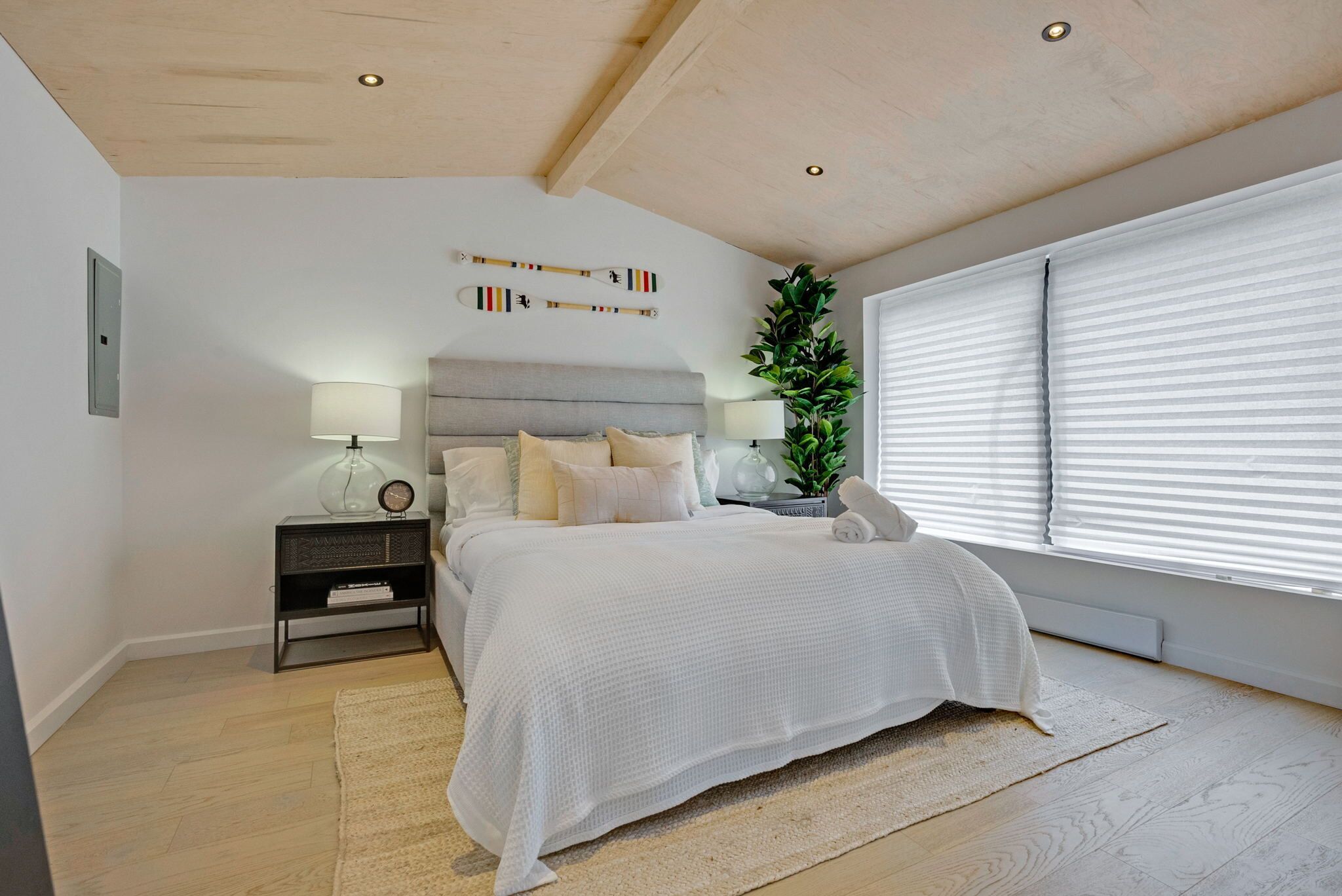 Un dormitorio con cama, mesitas de noche, lámpara, planta en maceta y ventana con persianas.