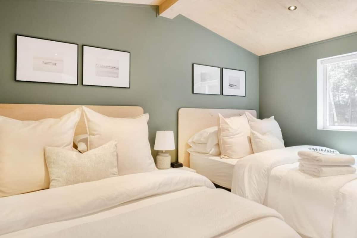 Habitación de hotel con dos camas, una mesita de noche con lámpara, cuadros en la pared y una ventana con vistas a árboles.