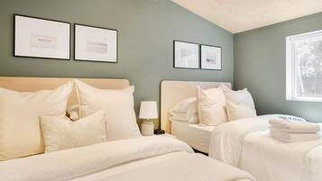 A hotel room with two beds, a nightstand with a lamp, framed pictures on the wall, and a window with a view of trees.