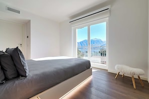 Ein modernes Schlafzimmer mit einem Bett, einem kleinen Hocker und einem großen Fenster mit Bergblick.