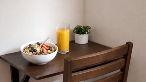 Ein Holztisch mit einer Schüssel Müsli und einem Glas Orangensaft, einer Topfpflanze und einem Stuhl mit grauem Sitzkissen.