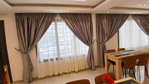 Un comedor moderno con una mesa y sillas de madera, ventanas amplias con cortinas y una máquina de café sobre un carrito.