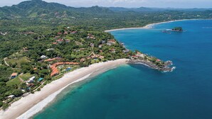Vista aérea de uma área costeira com praia, águas azuis cristalinas e um morro denso com edifícios.
