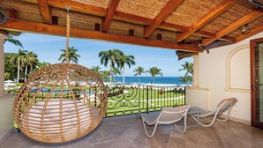 Varanda com cadeira suspensa, mesa e cadeiras, vista para uma praia com palmeiras e o oceano.