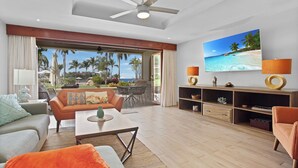 Uma sala de estar espaçosa com uma grande janela com vista para a praia, um ventilador de teto e uma televisão de tela plana na parede.