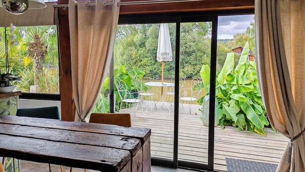 Een kamer met een houten tafel en stoelen, een groot raam met gordijnen en uitzicht op een terras met stoelen en planten.