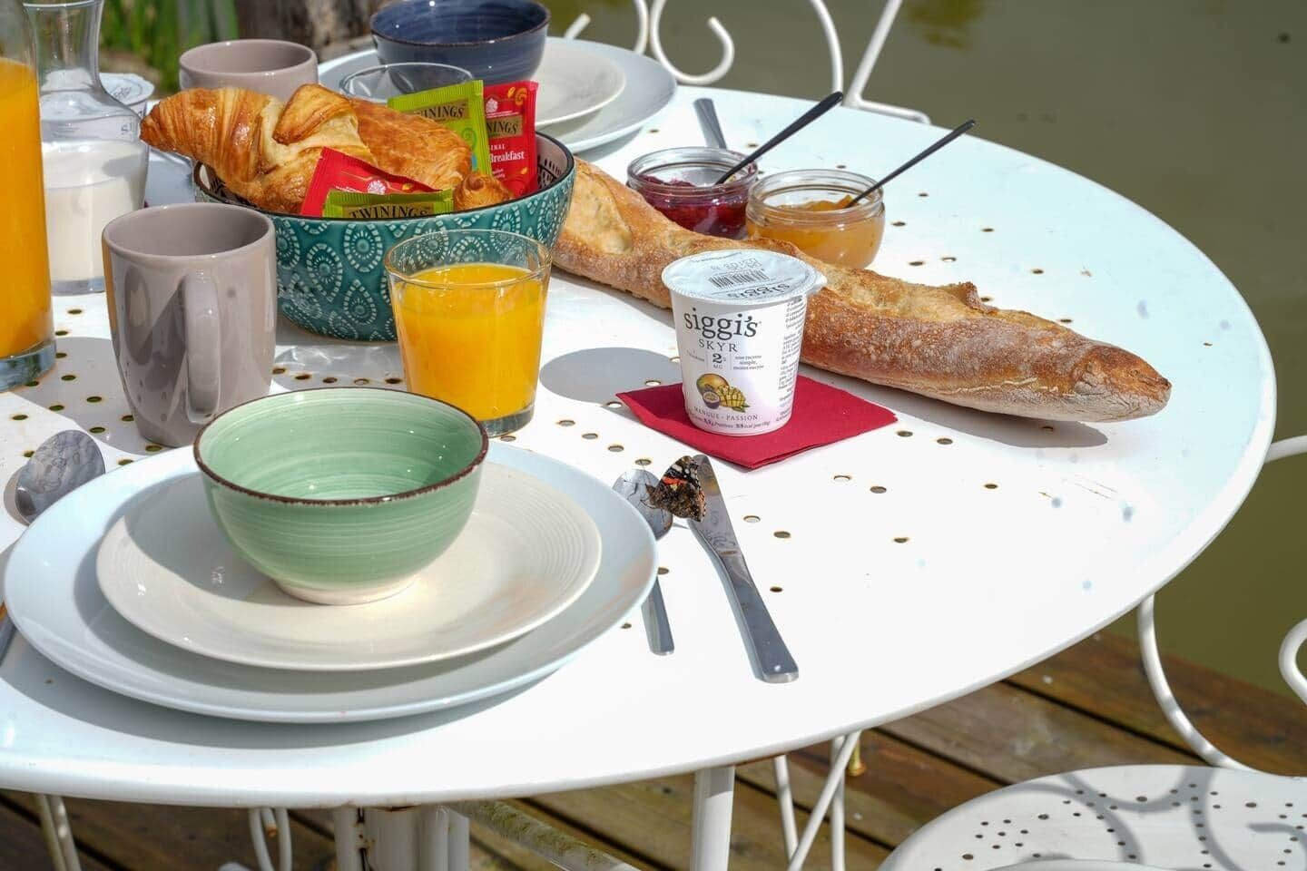 Una mesa de desayuno con cruasanes, un vaso de jugo de naranja, un tazón de yogur y una baguette.