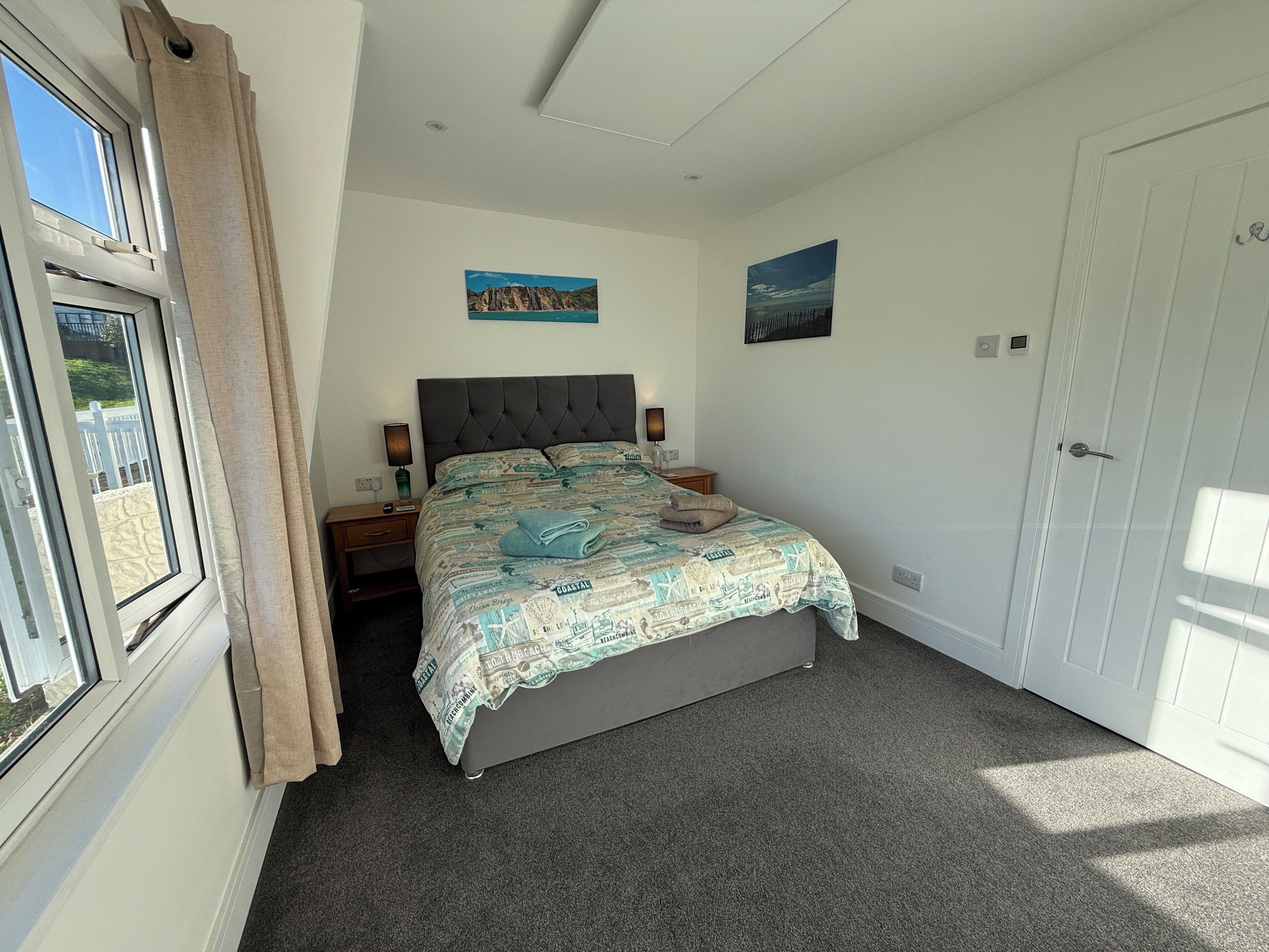 A bedroom with a bed, a nightstand, a lamp, a window with curtains, and a door.