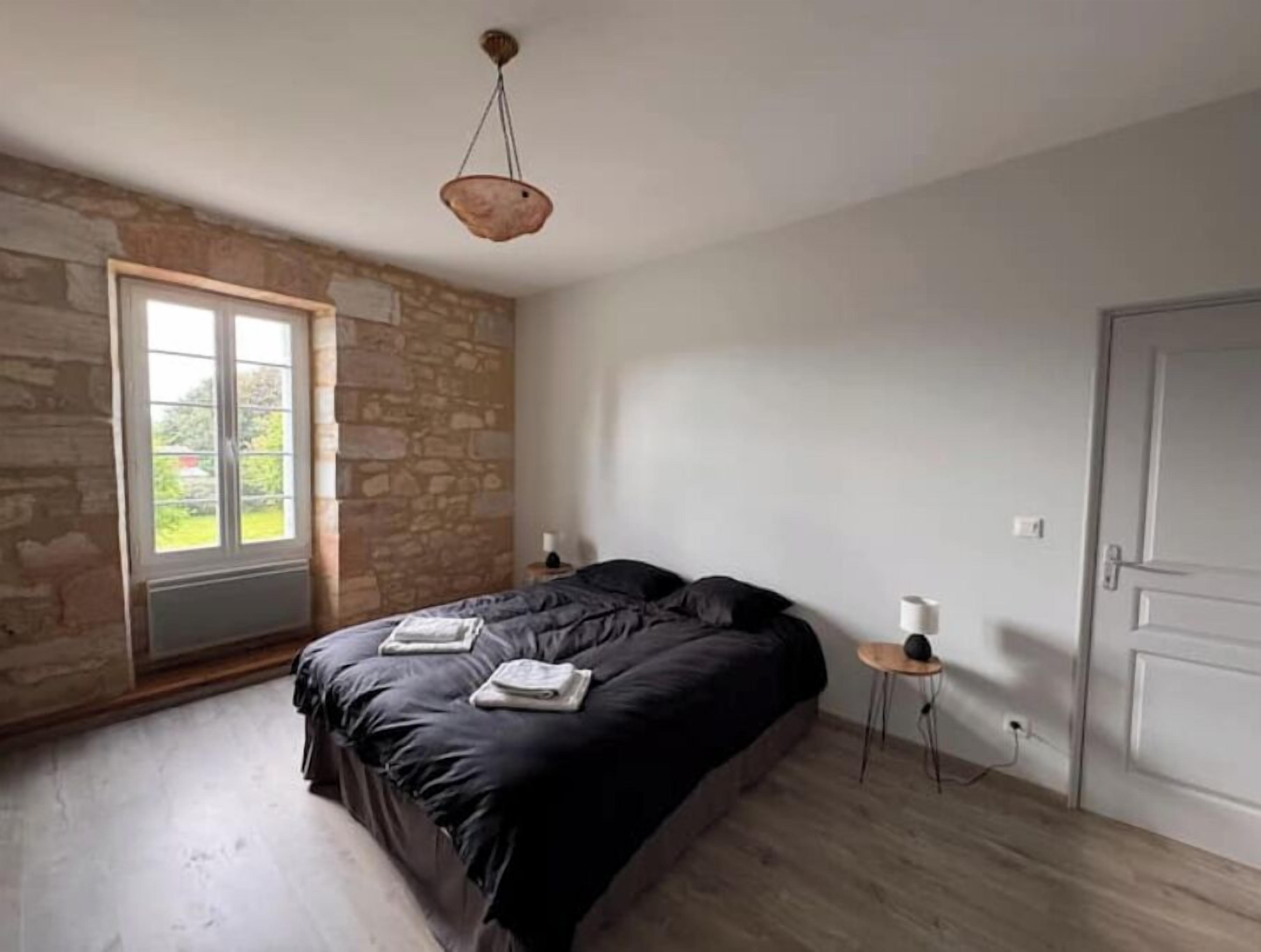 A bedroom with a stone wall, a large window, a bed with a dark cover, a bedside table with a lamp, and a door.