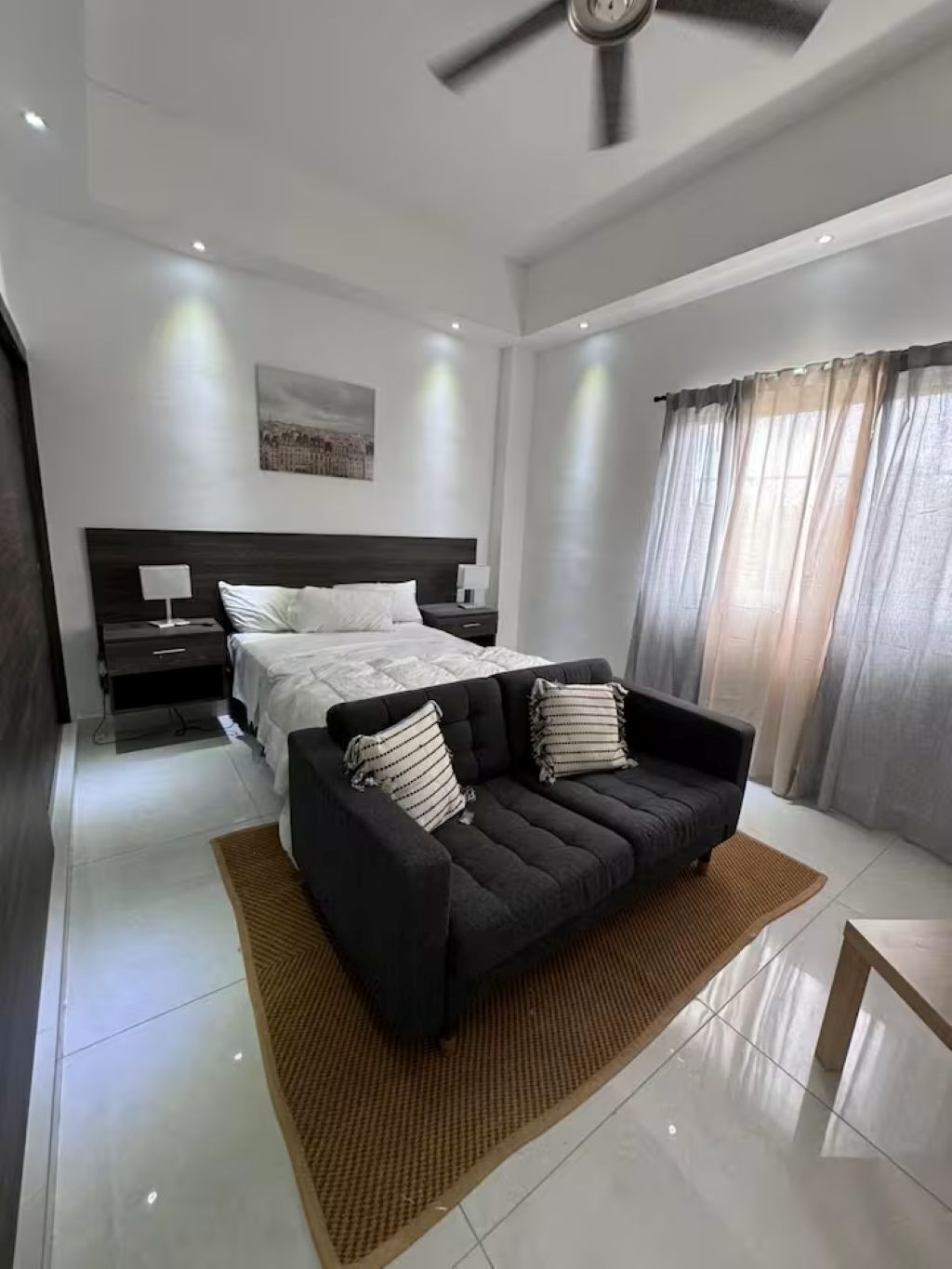 A modern bedroom with a large bed, a dark sofa, and a ceiling fan.
