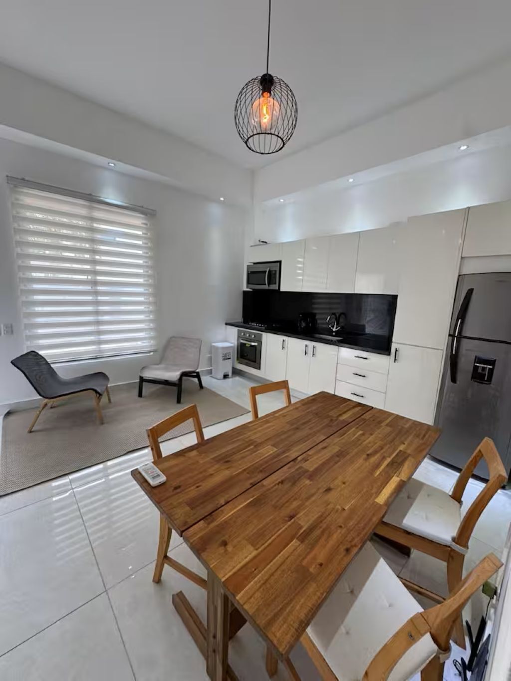 A modern kitchen with white cabinets, a wooden dining table, and a hanging light fixture.