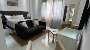 A modern hotel room with a bed, a black sofa, a beige armchair, a small table, and a ceiling fan.