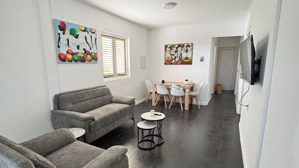 Un vaste espace de vie comprenant un canapé gris, une petite table ronde et une table à manger avec des chaises. Il y a deux fenêtres équipées de stores, un téléviseur à écran plat fixé au mur et des œuvres d’art accrochées aux murs.