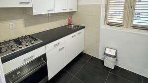 Une cuisine avec des meubles blancs, un plan de travail noir, une cuisinière à gaz, un four et une fenêtre avec des stores.