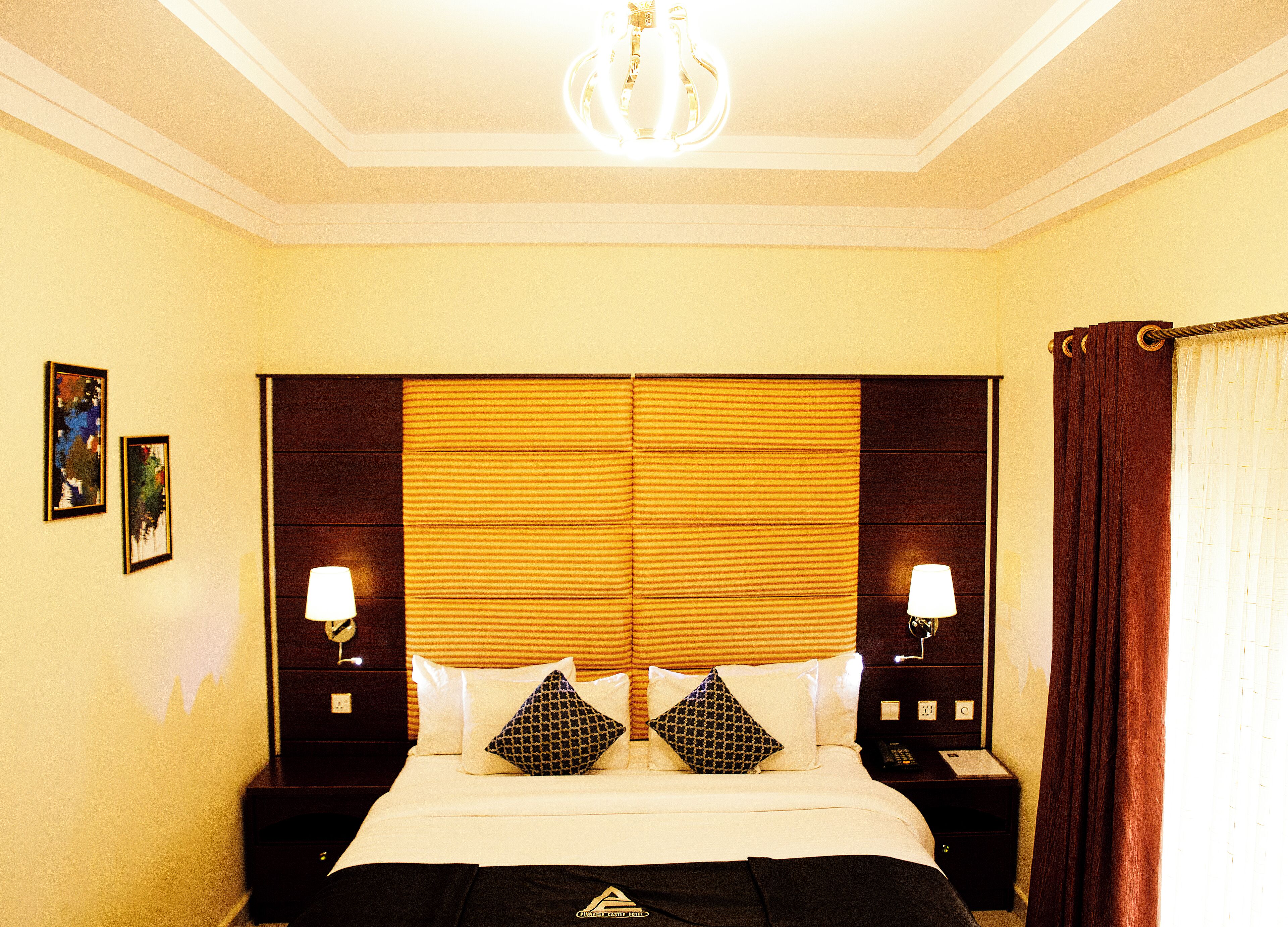 A hotel room with a large bed, two bedside tables with lamps, a chandelier, and a window with curtains.