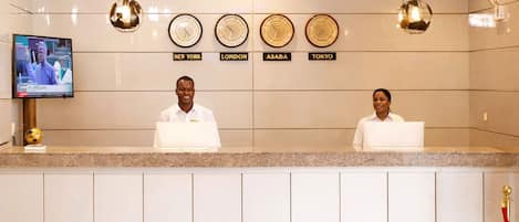 Un mostrador de recepción de hotel con relojes que marcan diferentes husos horarios, una pantalla de televisión y un piso de mármol.