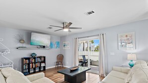 Ein Wohnzimmer mit einer Couch, Couchtisch, Bücherregal, Fernseher, Deckenventilator und Kunstwerken an den Wänden.