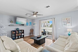 A living room with a sofa, coffee table, bookshelf, TV, ceiling fan, and artwork on the walls. - 🌴Walk to Beach + Community Pool | Summer Lake 23🌴 (Miramar Beach)