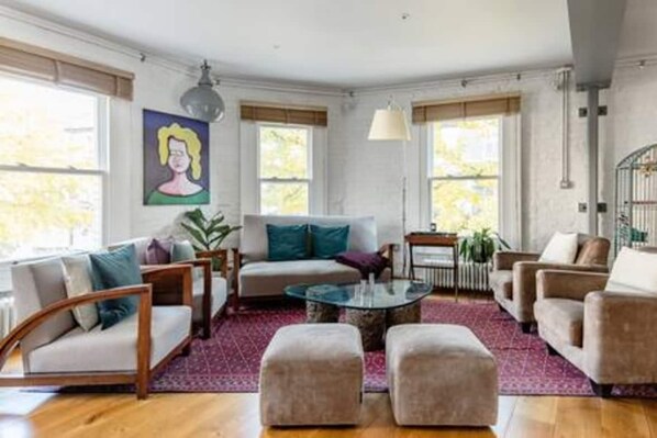 A modern living room with a sofa, armchairs, ottomans, and a coffee table. There are large windows with blinds, a painting on the wall, and a hanging light fixture. - Experience London in amazing reconverted Gin house (London)