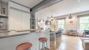 Cozinha moderna com armários brancos, uma ilha central com banquetas, uma área de refeições com cadeiras e vista para o exterior por grandes janelas.