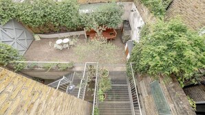 Um pátio externo com mesa e cadeiras de madeira, cercado por paredes de tijolo e vegetação.